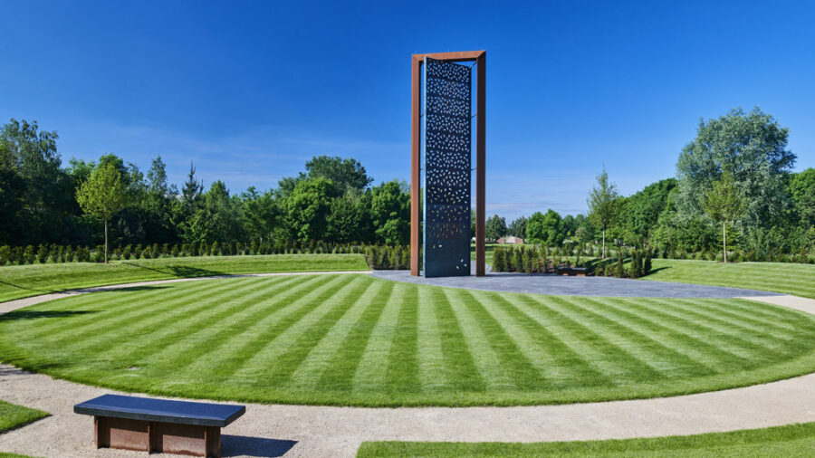 Tall bronze and stone police memorial with perforated brass screen, leaf‑shaped apertures, surrounding trees, and reflective open lawn.